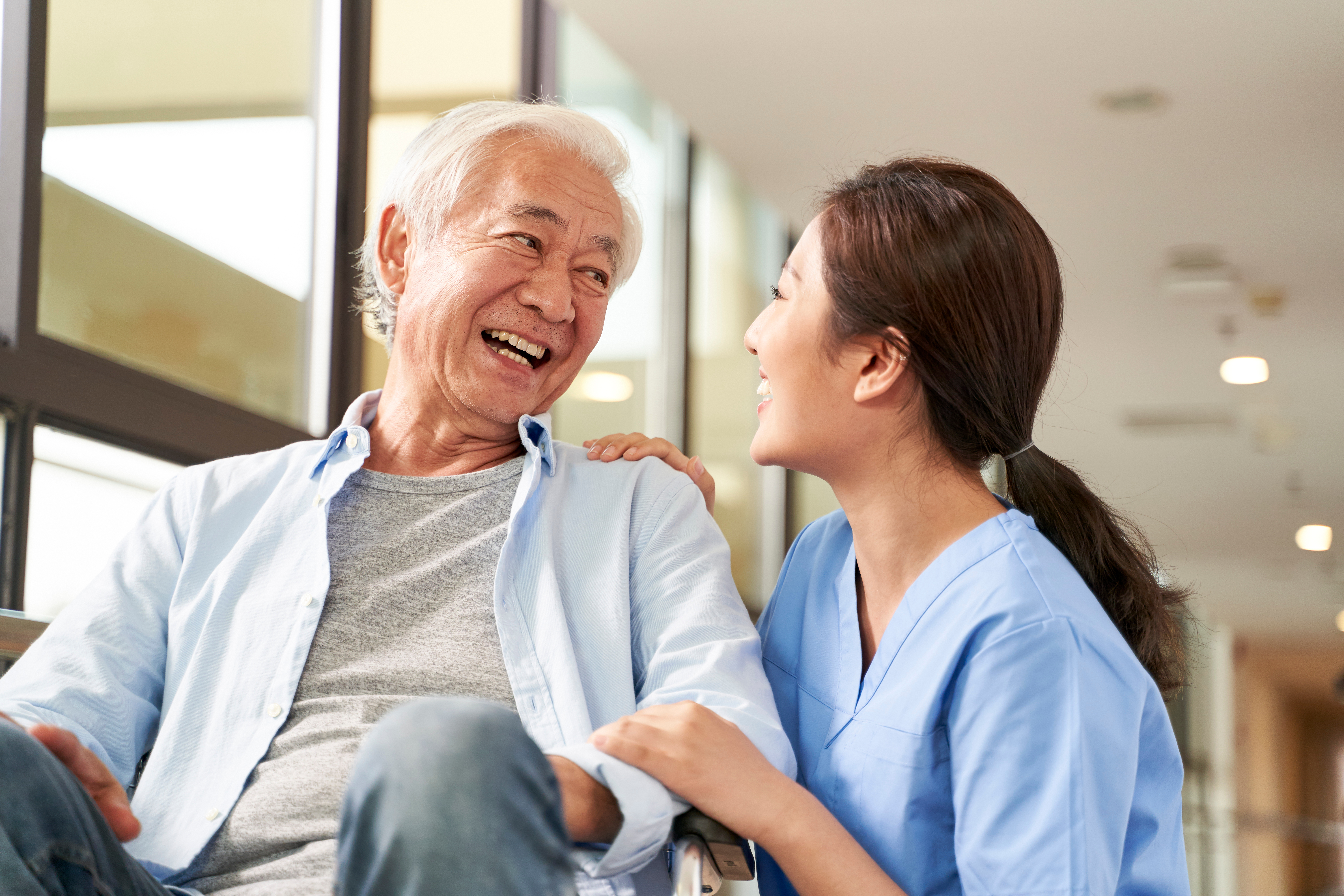 young friendly female caregiver talking chatting to happy senior man in hallway of nursing home